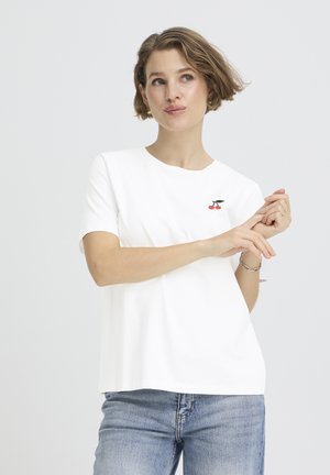 Femme aux cheveux courts portant un t-shirt blanc avec un logo cerise sur la poitrine et un jean bleu clair, ajustant des bagues à ses doigts.