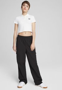 Biely skrátený polokošeľ s golierom a čiernym logom v kombinácii s čiernymi cargo nohavicami. Model má na sebe biele tenisky s hnedými podrážkami.