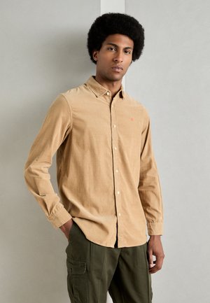 Tan corduroy shirt with long sleeves, button-down collar, and a small red logo. Worn with olive green cargo pants in a neutral background.