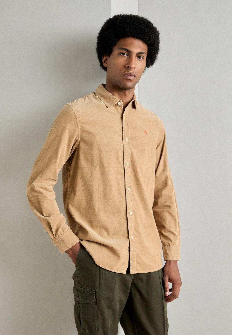 Camisa de pana marrón con mangas largas, cuello abotonado y un pequeño logo rojo. Llevada con pantalones cargo verde oliva sobre un fondo neutral.