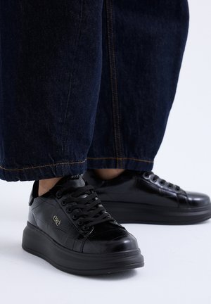 Zapatillas negras de plataforma con diseño texturizado, usadas con vaqueros anchos azul oscuro sobre fondo blanco.