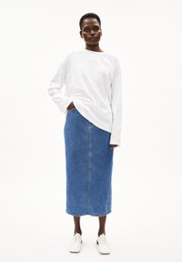 Camisa blanca de manga larga con un logo dorado, combinada con una falda midi de denim azul de cintura alta y zapatos blancos. Diseño simple y ajuste casual.