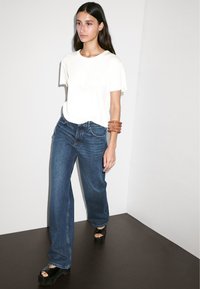 T-shirt bianca a maniche corte abbinata a jeans larghi blu scuro, bracciale marrone al polso e sandali platform neri. Tessuto morbido e vestibilità rilassata.