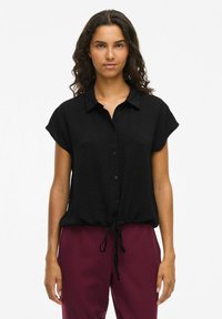 Črna kratkosleeve bluzica z gumbi na sprednji strani in zavezanjem na robu. Lahka tkanina z blagim teksturiranim vzorcem. V kombinaciji z burgundskimi hlačami.