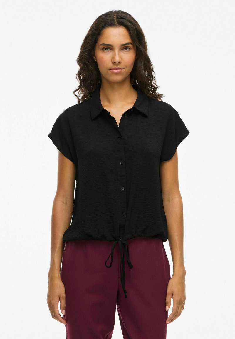 Črna kratkosleeve bluzica z gumbi na sprednji strani in zavezanjem na robu. Lahka tkanina z blagim teksturiranim vzorcem. V kombinaciji z burgundskimi hlačami.