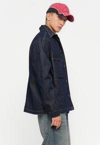 Jonge man met een rode en zwarte pet, een donkerblauwe spijkerjas met knoopzakken en lichtgewassen blauwe jeans, staande in zijaanzicht.