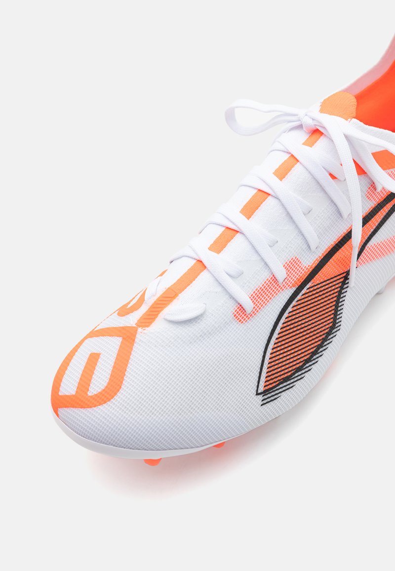 Chaussure de soccer blanche avec des accents orange, comportant un dessus texturé, un système de laçage et des semelles à crampons pour une meilleure traction. Le design inclut des détails noirs.