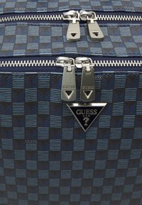 Mala de viagem preta e azul com fechos com zip, acessórios prateados e um detalhe de logotipo triangular. Tecido texturizado com design em padrão de tabuleiro de xadrez.