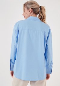 Light blue, long-sleeve shirt in a relaxed fit, featuring a traditional collar, back pleats, and cuffs with slits for adjusting sleeve length.