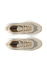 Beige atletiske sneakers med mesh-paneler, rund tå, tyk hvid sål og flade snørebånd. Har brandteksten synlig indeni.