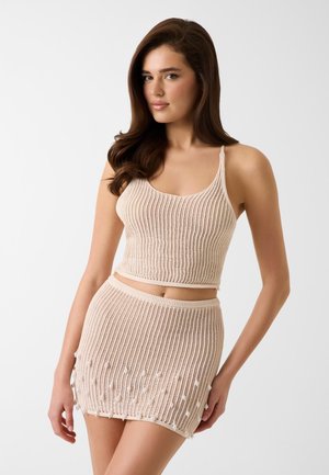 Kobieta z długimi brązowymi włosami, ubrana w beżowy dziergany krótki top oraz pasującą do niego mini spódnicę z drobnymi ozdobami z koralików na krawędzi spódnicy.