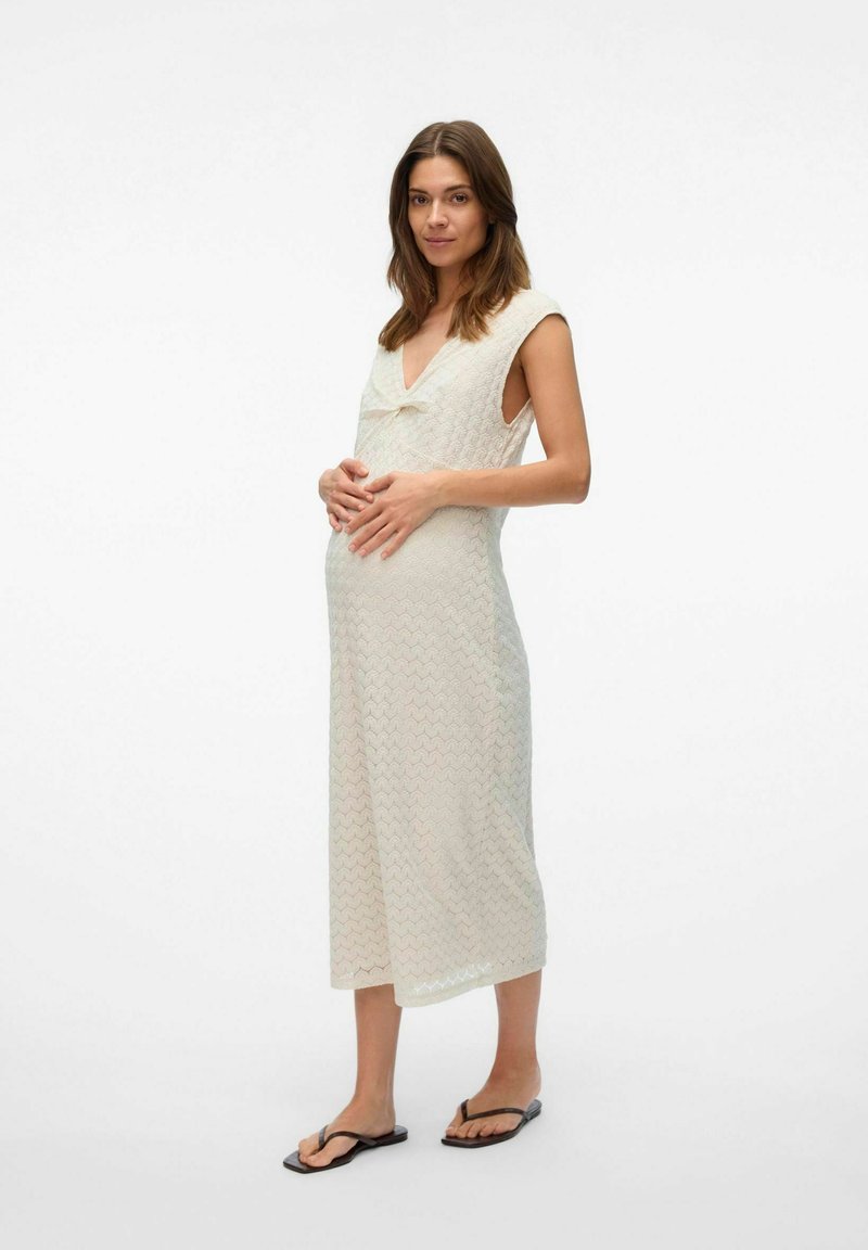 Robe tricotée légère de couleur crème avec un décolleté en V, des manches courtes et un motif en zigzag ; portée par un modèle enceinte avec des sandales plates.
