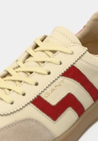 Baskets en cuir beige avec des accents rouges et blancs, dotées d'un bout texturé, de lacets jaunes et d'un logo GANT sur le côté.