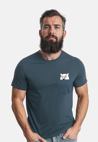 Behaarde man met een serieuze uitdrukking die een blauw T-shirt draagt met een klein wit cartoonbeertje dat uit de borstzak kijkt.