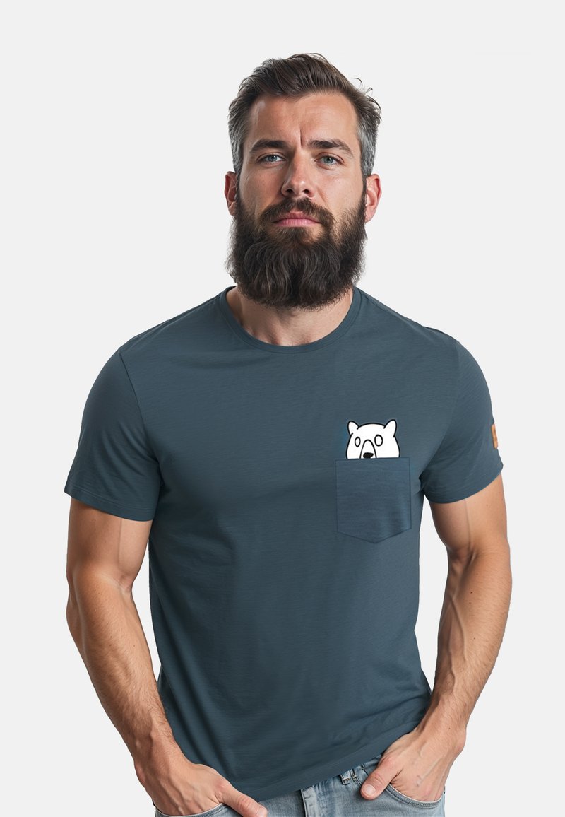 Behaarde man met een serieuze uitdrukking die een blauw T-shirt draagt met een klein wit cartoonbeertje dat uit de borstzak kijkt.