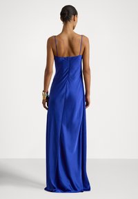 Femeie cu părul prins în coc, purtând o rochie lungă până la podea, din satin albastru regal, cu bretele subțiri, stând cu fața spre spate, pe un fundal simplu.