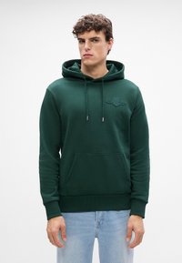 Felpa verde realizzata in misto cotone, con tasca a canguro, cappuccio regolabile con stringhe e logo ricamato sul petto. Tessuto morbido.