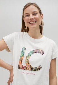Camiseta de algodón blanca con mangas cortas que presenta un colorido diseño de logo "LC" hecho de ilustraciones a lápiz. Texto debajo que dice "LOLA CASADEMUNT."