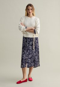 Strickpullover in Creme mit Kabelmuster, kombiniert mit einem marineblauen Rock mit weißen Blattmotiven und roten Ballerinas. Texturierte Lagen sind sichtbar.