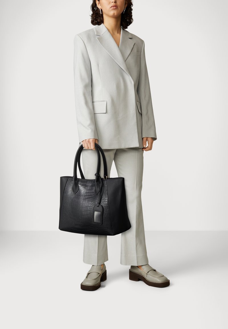 Personne portant un costume gris clair et des mocassins beige tenant un grand sac cabas en cuir noir texturé avec des poignées et une étiquette.
