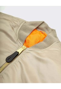 Beige bomber jacket featuring a ribbed collar, gold zipper closure, and an orange inner lining. Smooth texture with distinctive hardware detail.