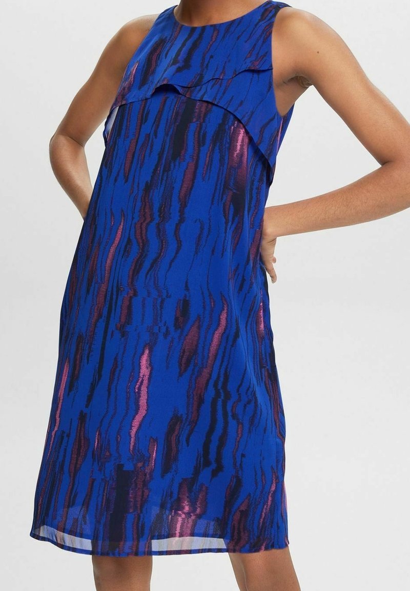 Robe sans manches longueur genou avec des motifs abstraits de bandes verticales bleues, noires et roses et un détail superposé au niveau de la poitrine portée par une personne debout.