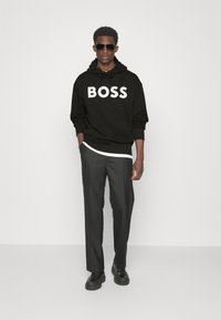 Felpa nera con la scritta "BOSS" in lettere bianche, abbinata a pantaloni scuri e scarpe nere. Il modello indossa occhiali da sole e ha le mani in tasca.