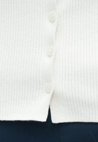 Bela rebrasta jopica s petimi belimi gumbi, ravnim robom in mehko teksturo. Na tkanini so subtilne navpične črte.