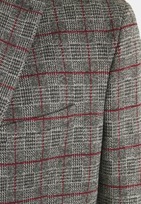 Grijze geruite blazer van een gestructureerde stof. Heeft een rood rasterpatroon en zwarte accenten. Toont naaddetail bij de schouder.