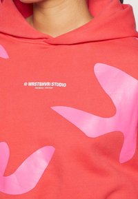 Sudadera de color coral con diseños asimétricos de pájaros en tono rosa. Presenta un logo en el frente, textura suave y capucha con cordones.