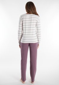 Woman standing barefoot facing away, wearing a long-sleeve white top with thin horizontal stripes and solid mauve pants.