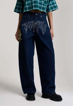 Persona vistiendo jeans de pierna ancha en azul oscuro con texto en estilo grafiti blanco que dice "Fearing" y bordado floral en el muslo izquierdo.