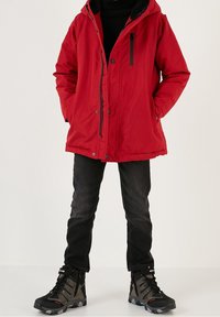 Punainen topattu takki, jossa on huppu, vetoketju ja painonapit sekä mustia yksityiskohtia. Yhdistettynä mustiin farkkuihin ja tukevan näköisiin mustiin vaelluskenkiin.