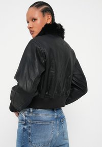 Osoba z warkoczami, nosząca czarną kurtkę z eko-skóry z futrzanym kołnierzem i niebieskie dżinsy, lekko zwrócona na bok.