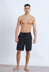 Άνδρας χωρίς πουκάμισο στέκεται ξυπόλυτος σε γκρι δάπεδο, φοράει μαύρο μαγιό μέχρι το γόνατο με κορδόνι στη μέση, μπροστά σε απλό ανοιχτό φόντο.