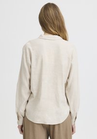 Femme aux longs cheveux châtain clair portant une chemise ample à manches longues beige et un pantalon marron, vue de dos sur un fond uni.