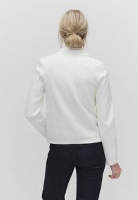 Bílá zipová bunda se strukturovaným designem, hladkou texturou a dlouhými rukávy. Nosená s tmavými džíny, pohled zezadu.