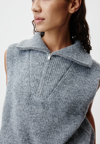 Frau trägt einen weichen, strukturierten ärmellosen grauen Strickpullover mit einem halben Reißverschluss-Kragen und sitzt leicht nach links gerichtet vor einem schlichten Hintergrund.