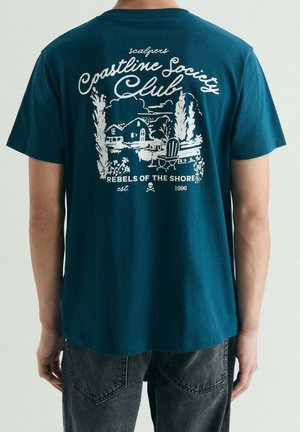 Persona con camiseta de color teal oscuro con un gráfico blanco de una casa, árboles, silla y texto "Coastline Society Club" y "Rebels of the Shore".