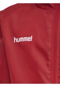 Giacca rossa con una texture liscia, dotata di cappuccio, zip frontale e un logo "hummel" bianco sul petto.