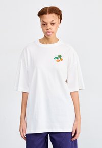 Balta per didelė medvilninė marškinė, su žaliais ir oranžiniais grafikos akcentais priekyje, įskaitant Nike logotipą ir vaisių motyvus.