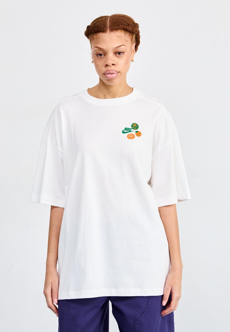 Camiseta de algodón blanca de gran tamaño con detalles gráficos en verde y naranja en la parte frontal, que incluyen un logo de Nike y diseños de frutas.