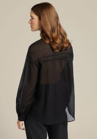 Blusa nera trasparente con maniche lunghe, dettagli testurizzati sul retro e orlo arrotondato. Il tessuto leggero conferisce un aspetto morbido e fluido.
