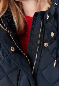 Primo piano di una persona bionda che indossa un maglione rosso e una giacca nera trapuntata parzialmente chiusa, con cerniere e bottoni dorati.