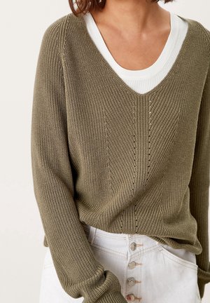 Kvinde iført olivengrøn strikket V-hals sweater over hvid top, kombineret med højtaljede hvide bukser med knapper, vist fra hals til hofter.