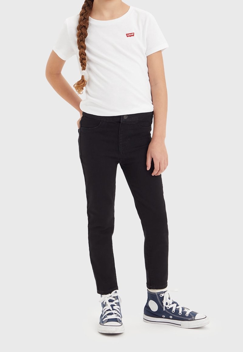 Niño con cabello largo trenzado, vistiendo una camiseta blanca de Levi's, jeans negros y zapatillas altas azules, de pie contra un fondo simple.