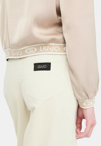 Blusa beige chiaro con maniche a sbuffo e elastico. Pantaloni in corduroy crema con un'etichetta nera sul taschino posteriore. Texture liscia.