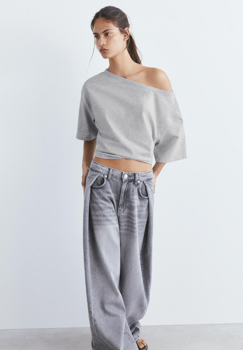 Jonge vrouw staat met handen in de zakken, draagt een losse grijze off-shoulder cropped top en losse, vervaagde grijze spijkerbroek tegen een effen achtergrond.