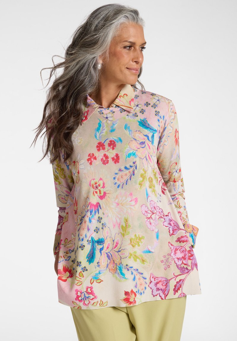 Donna con capelli grigi che indossa una maglia a maniche lunghe con motivo floreale pastello e colletto, pantaloni giallo chiaro, mani in tasca, guarda verso destra.