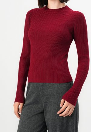 Femme portant un pull bordeaux à manches longues côtelé ajusté et un pantalon gris à chevrons, debout devant un fond blanc.
